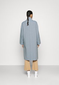 Donna con capelli intrecciati che indossa un lungo cappotto blu, pantaloni beige a gamba larga e stivaletti bianchi alla caviglia, in piedi di fronte a una parete chiara e semplice.