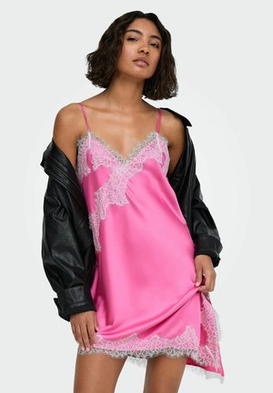 Femme portant une robe nuisette en satin rose avec une bordure en dentelle blanche et une veste en cuir noire drapée sur ses épaules.