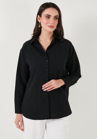 Black button-up shirt with a ribbed texture, featuring a collar and long sleeves, paired with light-colored pants.