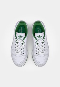 Bílé kožené tenisky se zelenými akcenty, vybavené perforovanou špičkou, šněrováním a polstrovaným límcem. Logo Adidas na jazyku a stélce.