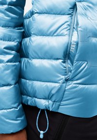 Lichtblauwe pufferjack met gequiltte delen, een zijzak met rits en een verstelbare trekkoord onderkant. Gladde, glanzende afwerking.
