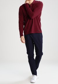 Burgundowa koszulka polo z długim rękawem, ciemnogranatowe spodnie i białe sneakersy. Koszulka polo ma kołnierzyk i zapięcie na guziki; spodnie mają dopasowany krój.