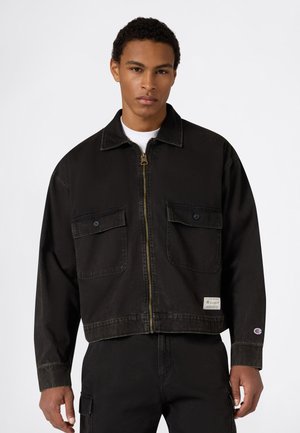 Joven con chaqueta negra con cremallera, dos bolsillos en el pecho con botones y pantalones negros, sobre un fondo liso.