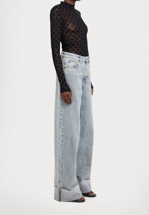 Osoba ubrana w czarną, wzorzystą, przezroczystą bluzkę z długim rękawem, wysokie spodnie z szerokimi nogawkami w jasnym odcieniu dżinsu oraz czarne sandały na obcasie z odkrytymi palcami.