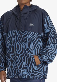 Tamno plavi pulover jakna s kapuljačom, s valovitim svijetloplavim uzorkom, džepom na zip, i elastičnim manžetama. Lagani materijal.