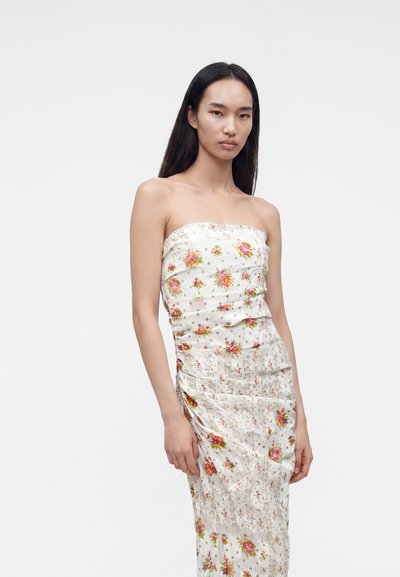 Jeune femme aux longs cheveux noirs portant une robe blanche sans bretelles avec des motifs floraux colorés, debout devant un fond clair uni.