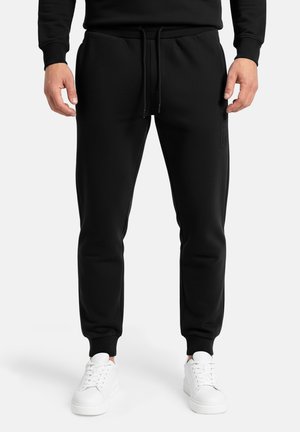 Uomo che indossa pantaloni da jogging neri con coulisse in vita e scarpe da ginnastica bianche, in piedi davanti a uno sfondo chiaro e uniforme.