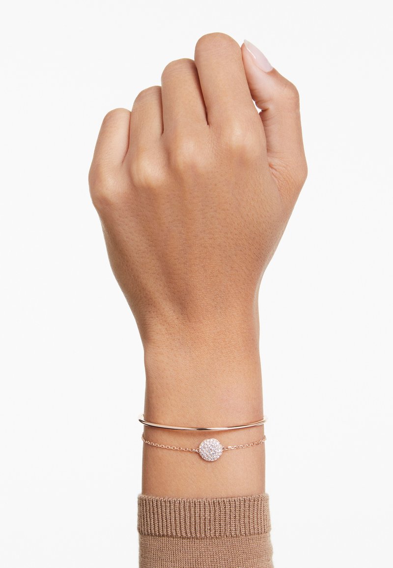 Pulseras de oro rosa en una muñeca; una es un brazalete liso, la otra presenta un charm redondo con piedras claras y brillantes. El tono de piel es medio.