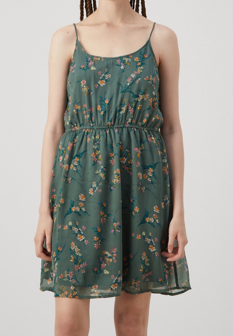 Robe verte sans manches avec fines bretelles, taille cintrée et petit imprimé floral coloré portée par une personne debout sur fond uni.