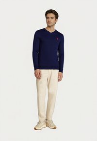 Nuori mies seisoo päällään tummansininen V-kaula-aukkoinen villapaita, vaaleat beige housut ja beige tennarit yksinkertaisella valkoisella taustalla.