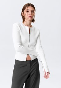 Pull blanc côtelé à fermeture éclair avec un col rond et une coupe ajustée, présentant des manches allongées et un accent zipper à l'ourlet.