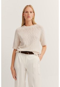Helles beiges Strickpullover mit kurzen Ärmeln und rundem Ausschnitt, kombiniert mit cremefarbener Hose und einem braunen Ledergürtel. Dezent strukturierte Oberfläche sichtbar.