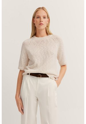 Helles beiges Strickpullover mit kurzen Ärmeln und rundem Ausschnitt, kombiniert mit cremefarbener Hose und einem braunen Ledergürtel. Dezent strukturierte Oberfläche sichtbar.
