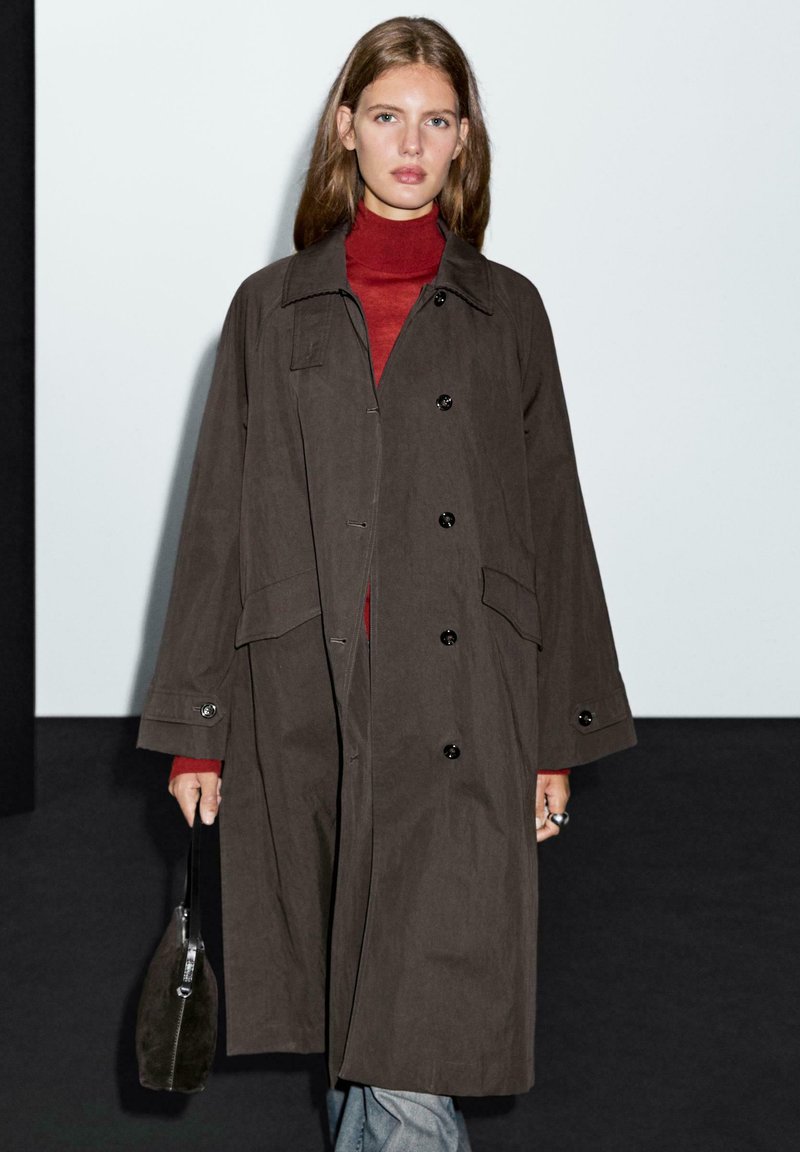 Hnědý oversized kabát s límcem, zapínáním na knoflíky a bočními kapsami, nošený přes červený top. Modelka drží malou černou kabelku.