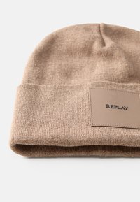 Brun strikket beanie med en foldbar kant, prydet med et rektangulært beige lædermærke med teksten "REPLAY." Blød tekstur, enkelt design.