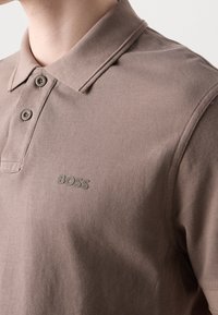 Gros plan sur une personne portant un polo taupe avec deux boutons et le logo BOSS en relief sur le côté gauche de la poitrine.