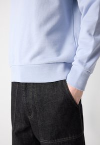 Personne portant un sweat-shirt bleu clair aux manches retroussées et un jean en denim noir, la main posée de manière décontractée dans la poche du jean.