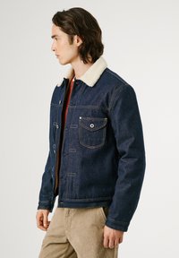 Chaqueta de denim con cuello de borreguito, color azul oscuro, con bolsillos tipo botón y costuras en contraste. Diseño simple, mangas de longitud media.