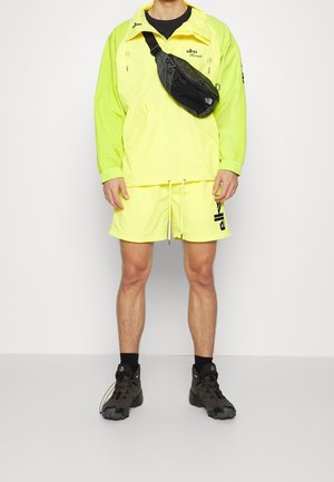 Veste coupe-vent jaune fluo et shorts assortis, sac bandoulière noir et chaussures de sport noires. Présente une taille élastique et des accents de logo.