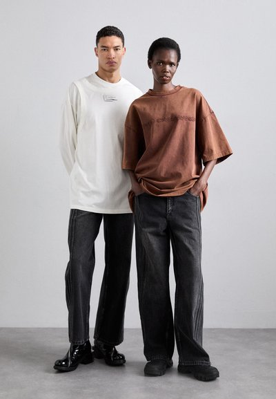 Two models stand side by side. The male wears a white long-sleeve shirt and black jeans. The female wears a brown oversized T-shirt and black jeans.