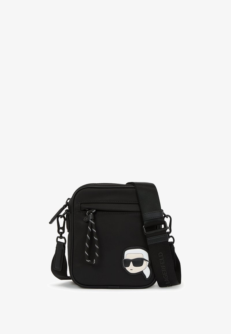 Čierna crossbody taška vyrobená z nylonu. Má hladkú textúru, zipsové uzavretie, nastaviteľný popruh a na prednej strane je nášivka s karikatúrou.