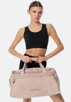 Športovo zdatná žena v čiernej športovej podprsenke drží veľkú béžovú športovú tašku Cardio Bunny s dvoma rukoväťami na bielom pozadí.