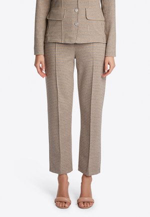 Vrouw draagt beige en bruine op maat gemaakte broek met een hondentandpatroon en bijpassend jasje met beige sandalen met open teen.