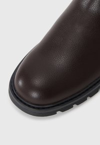 Botín de cuero marrón con textura suave, puntera redondeada y suela de goma negra contrastante, con un diseño de banda de rodadura resistente.