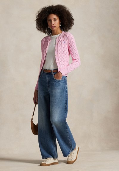 Moteris su garbanotais plaukais, dėvinti rožinį megztą kardiganą, baltą marškinėlį, platėjančias džinsines kelnes, baltus sportinius batelius ir laikanti rudos spalvos rankinę.