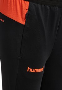 Black athletic pants with an orange mesh panel and zipper detail, featuring the white Hummel logo on the lower leg.