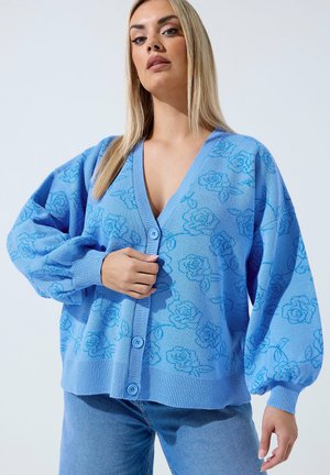 Femme blonde portant un cardigan bleu ample avec un motif de roses, boutonné devant, associé à un jean bleu sur un fond uni.