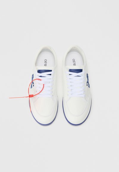 Fehér tornacipők kék díszítéssel, texturált anyaggal, lapos fűzőkkel és látható márkajellel a nyelvén. Tartalmaznak egy piros cipzáras részletet.