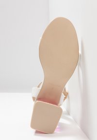 Semelle beige d'une sandale blanche pour femme avec talon bloc, taille 4, appuyée contre un mur et un sol blancs.