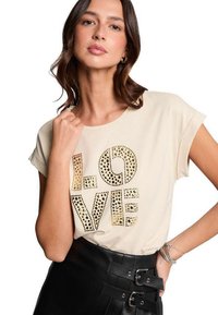 Crème t-shirt met het woord "LOVE" in gestructureerde, goudkleurige versieringen. Korte mouwen, ontspannen pasvorm, gecombineerd met een zwarte rok.