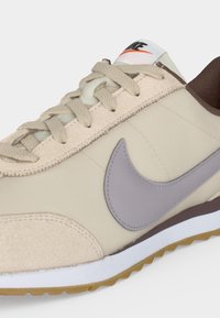 Μπεζ αθλητικό παπούτσι Nike με λεκέ μπλε ταπουέ, υφασμένα κορδόνια, λευκή ενδιάμεση σόλα, καφέ γιακά και εξωτερική σόλα από καουτσούκ, εμφανίζεται από κοντινή πλευρική γωνία.