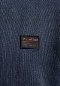 Țesătură texturată de un albastru închis, cu o etichetă neagră pe care se află "Petrol Ind" și "Denim Driven" în text portocaliu, pe un fundal țesut.