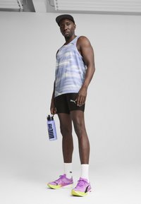 Lilla tie-dye tank top, sorte shorts med logo, lilla og gule atletiske sko, holder en gennemsigtig vandflaske med sort skrift.