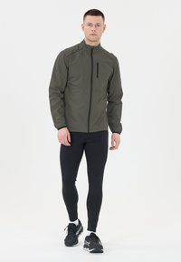 Man som går framåt iförd en grå zip-up jacka, svarta tights, vita strumpor och svarta löparskor mot en vit bakgrund.