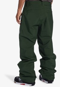 Green snowboarding pants with a loose fit, made of waterproof material, featuring rear pockets and a logo patch on the left hip.