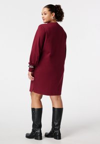 Bordeaux lange mouw jurk met zijsplitten. Glad textuur, relaxte pasvorm. Draag met kniehoge zwarte laarzen, met gespaccents.