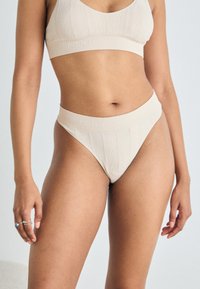 Beige kootud bralette ja sobivad kõrge vöökohtadega stringid logoga detailiga. Kangas on tekstuurne muster ja sile viimistlus.