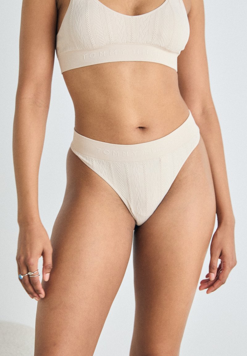 Beige kootud bralette ja sobivad kõrge vöökohtadega stringid logoga detailiga. Kangas on tekstuurne muster ja sile viimistlus.