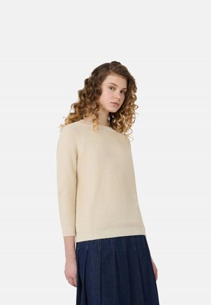 Giovane donna con capelli ricci che indossa un maglione beige lavorato a maglia e una gonna plissettata blu scuro, in piedi davanti a uno sfondo chiaro e uniforme.