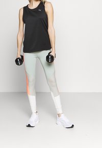 Černý nátělník bez rukávů, světle zelené legíny s oranžovými detaily a bílé tenisky. V obou rukou drží činky. Jednoduchý a funkční design.