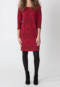 Czerwona sukienka do kolan z fakturowanym kwiatowym wzorem, rękawy do połowy przedramienia, zestawiona z czarnymi rajstopami i botkami za kostkę.