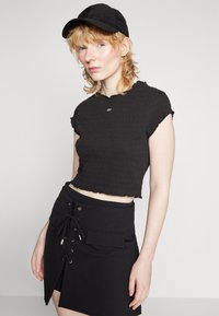 Sort smocked crop top med flæser langs kanten, parret med en sort miniskirt med front snøredetalje og lommer. Sort cap accessory.