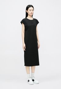 Fekete pamut ruha rövid karimás ujjakkal, kerek nyakkivágással és egyenes sziluettel. A mellrészen kis dombornyomott logó található.