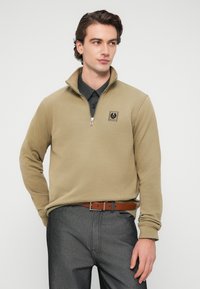 Μπεζ φούτερ με φερμουάρ και ψηλό γιακά, με μικρό λογότυπο στο στήθος. Φοριέται πάνω από σκούρο πουκάμισο με γιακά και γκρι παντελόνι.