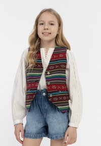 Mehrfarbige Weste mit rotem Grund, ausgestattet mit horizontalen Streifen und geometrischen Mustern, getragen über einer weißen, strukturierten Bluse und Denim-Shorts.