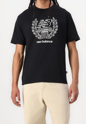 Person trägt ein schwarzes New Balance T-Shirt mit weißem Lorbeerkranz- und Säulenlogo, kombiniert mit beigen Hosen.